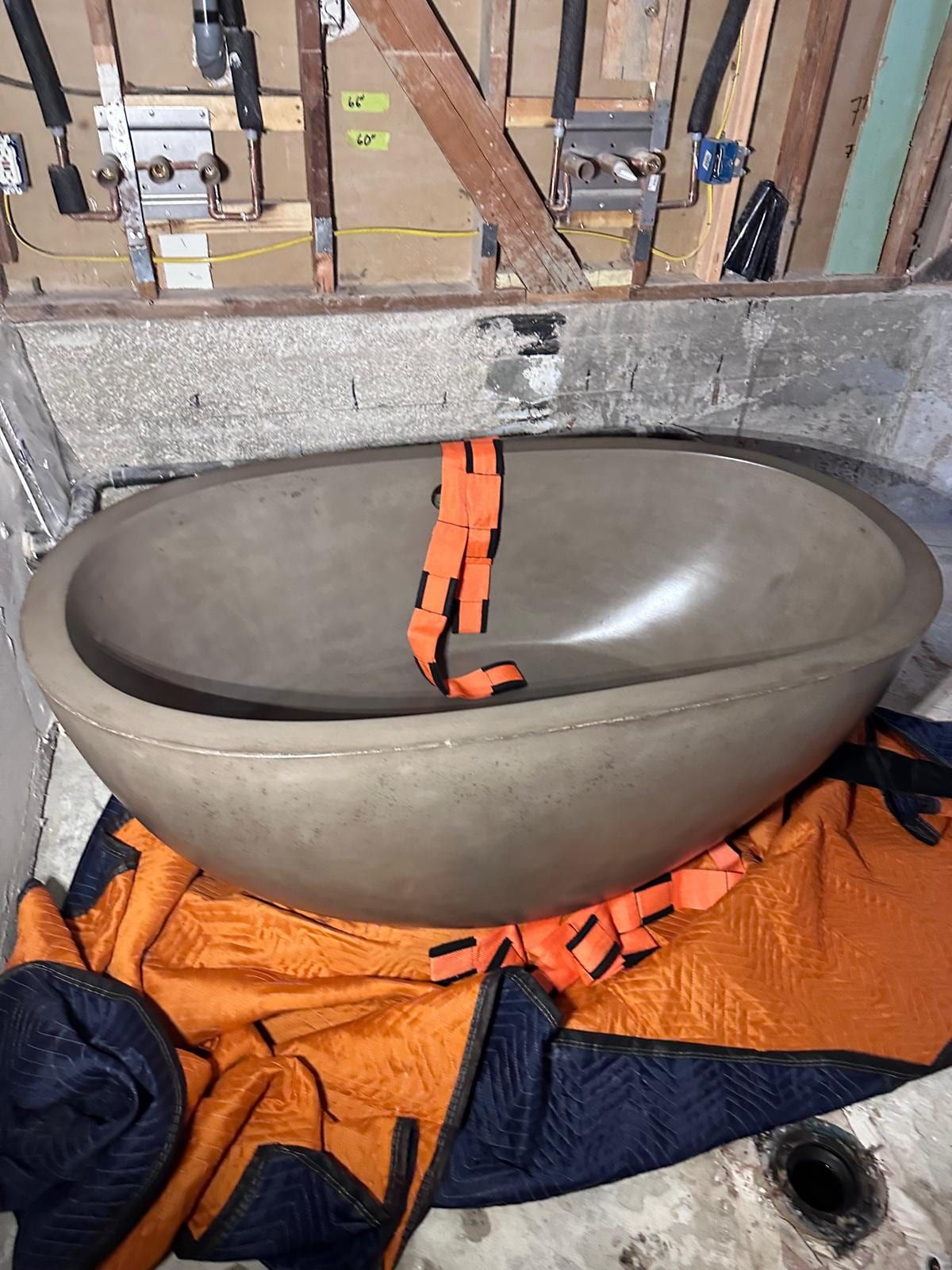 A concrete freestanding tub resting on moving blankets with its lifting straps, below an open wall with new valve rough-ins