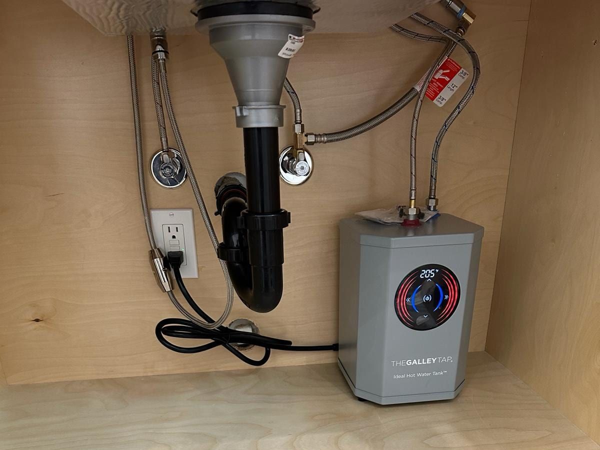 An under-sink instant hot water tank with a digital temperature display, connected beside the sink drain and supply valves