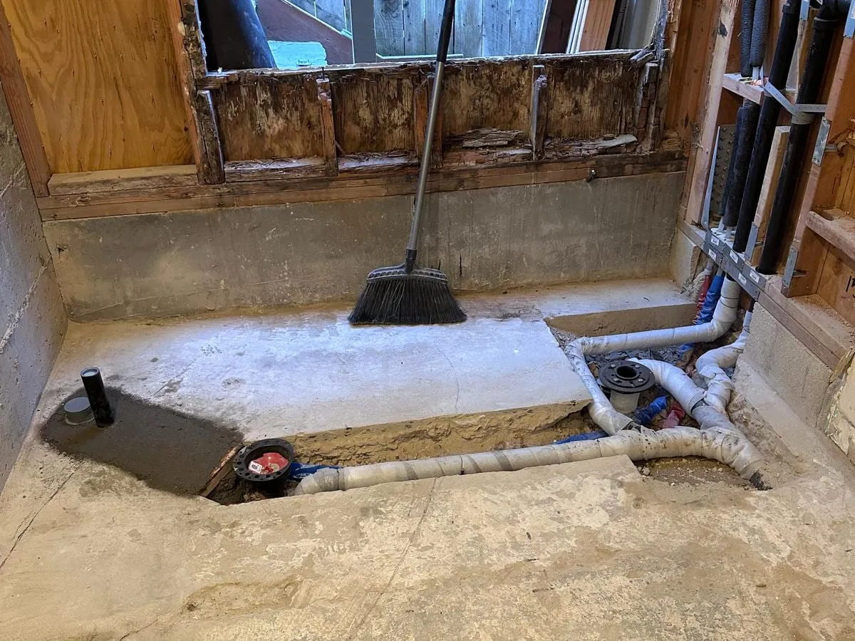A gutted bathroom with a section of the concrete slab cut open to show new drain lines and a capped toilet flange