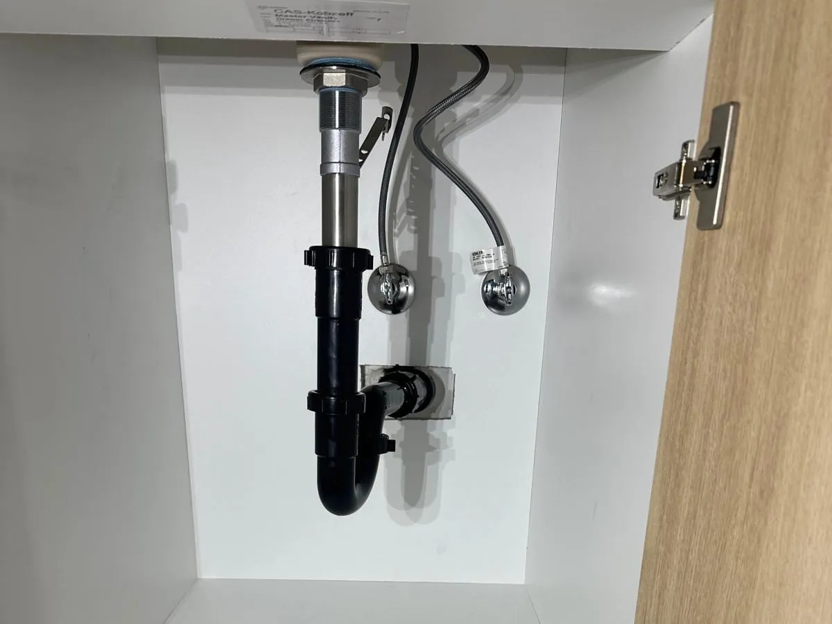 Under a bathroom vanity, a black drain trap, a chrome tailpiece and two supply valves inside a white cabinet